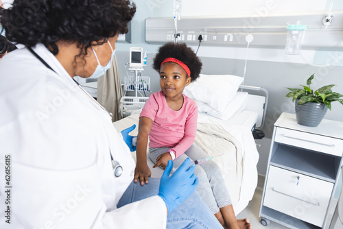 African american female doctor wearing face mask vaccinating girl patient at hospital