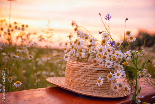 Fotografia, Obraz A straw hat and a bouquet of wild flowers on a wooden bench in a field at sunset on a summer evening