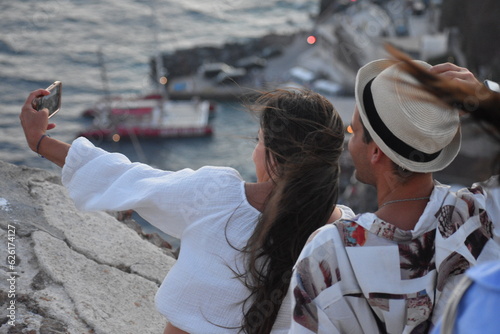 Happy Couple sitting on holiday taking selfie with Santorini island coastal panorama in Greece. Summer vacation, having Fun, Relax.