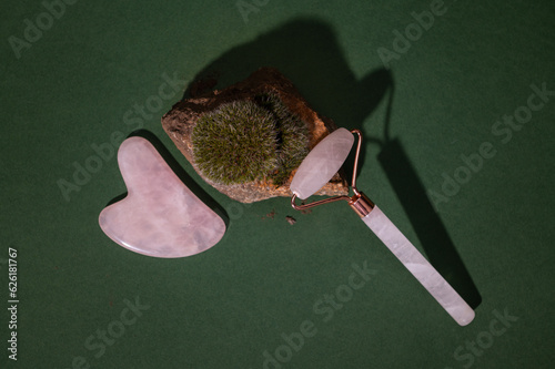 Flat lay. Massage tools - quartz stone roller massager and gua sha scraper on a rock with growing moss, green background