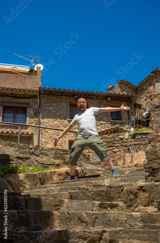 Bald man with half a beard smiling with open mouth, expressive eyes and arms outstretched to give a cheerful and funny surprise standing on a stone staircase in a rural village on a sunny day © sirbouman