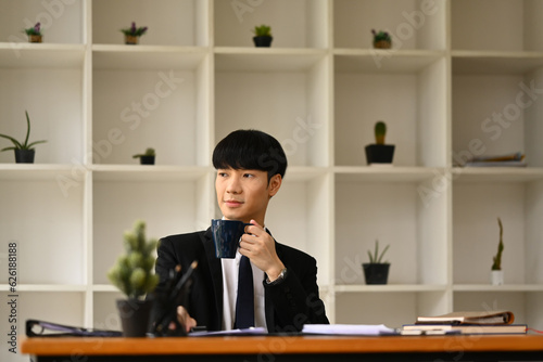 Thoughtful Asian male entrepreneur sitting at office desk and looking away, thinking of future career vision