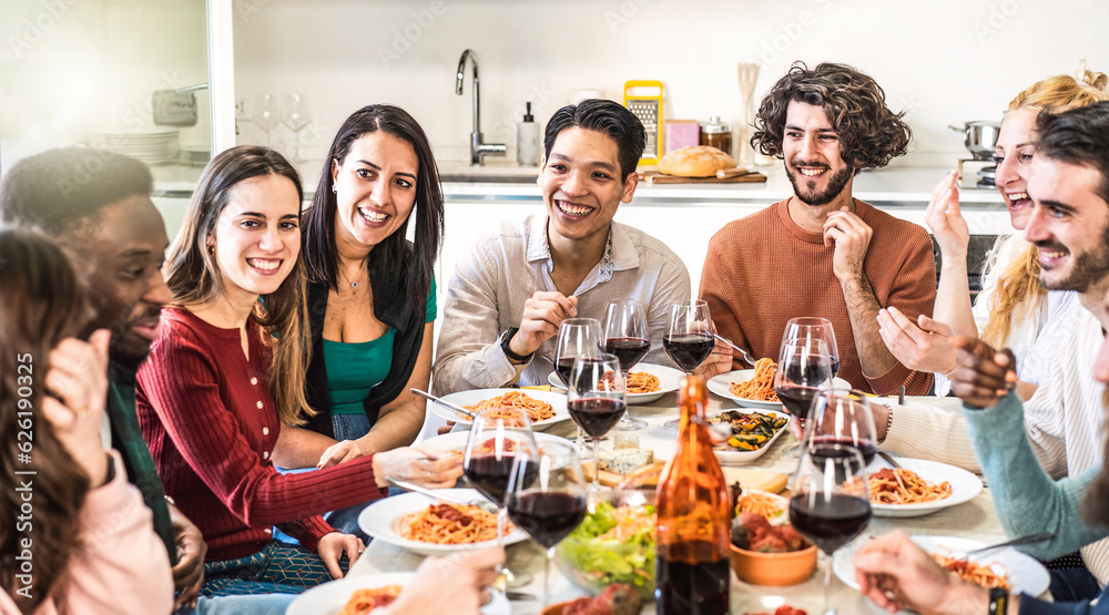 Happy multiethnic friends eating spaghetti or noodles with tomato sauce and drinking red wine at home during a amazing dinner party - Conviviality concept with bright warm filter