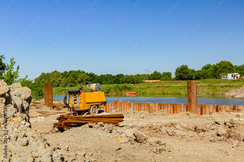 Bridge foundation made of metal piles on river coast, armature bundles and compressor placed at building site