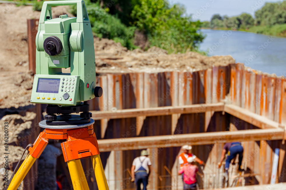 Total station on building site, in background is frame of metal piles for bridge foundation