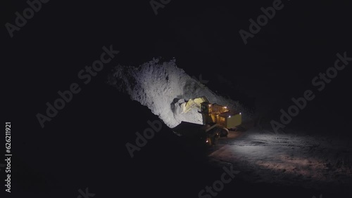 Escavators and dumper trucks work at night digging and transporting gold ore from open mine pit, slow motion