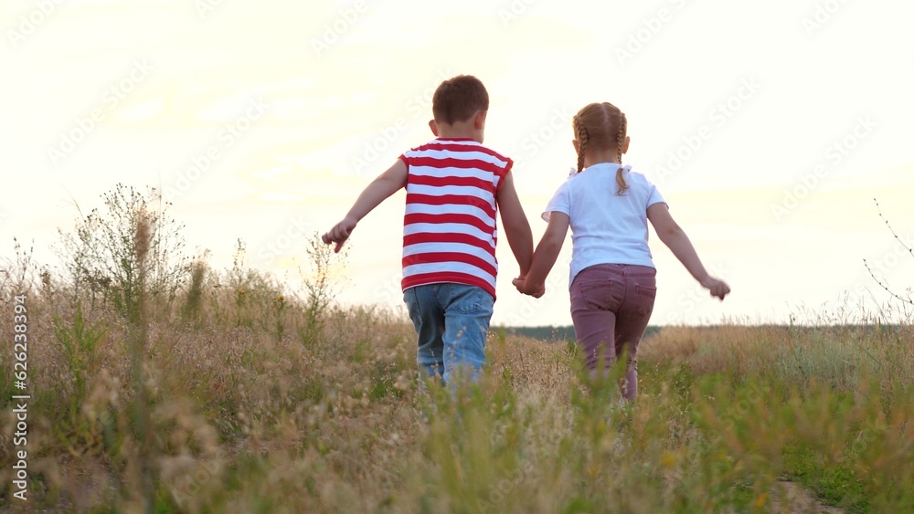 Cute playful boy and girl run joining hands along rural road with growing grass