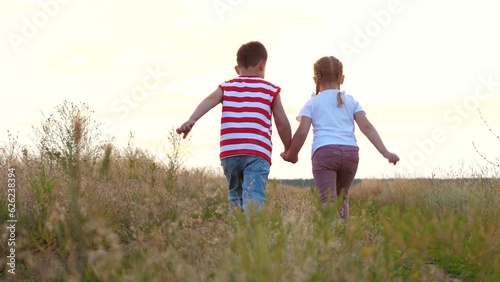 Cute playful boy and girl run joining hands along rural road with growing grass