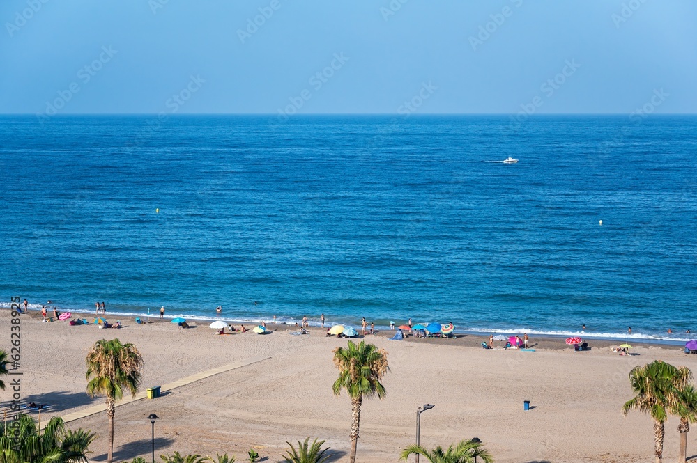 People enjoying a day at the beach, people with umbrellas on the seashore, others bathing, a boat sailing and palm trees on the sand