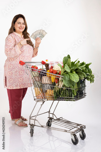 Photography Happy woman posing with a shopping cart full of groceries and holding money isolated on white background