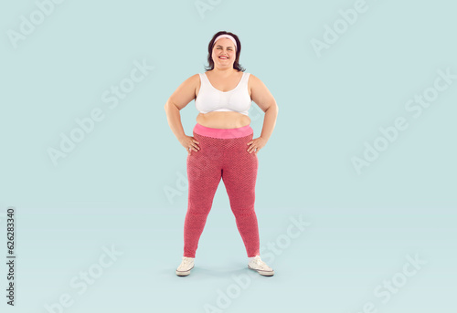 Happy funny young fat woman in sportswear looking at camera standing confidently with hands on the sides isolated on a studio blue background ready for sport fit exercises. Workout, gym concept.