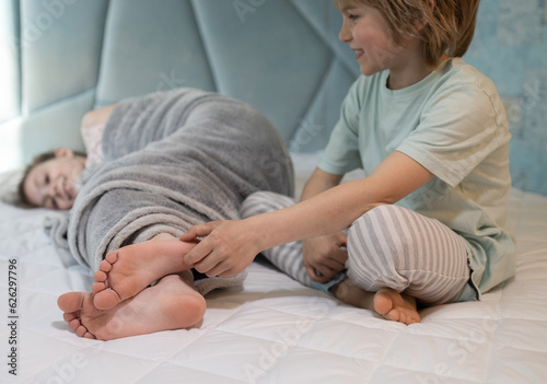 Photos cute boy, brother tickles the bare feet of the girl, his sister wrapped in a blanket lying on the bed at home