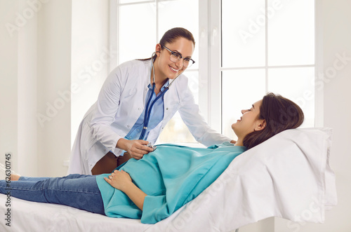 Friendly smiling gynecologist examining heartbeat in the abdomen of pregnant young woman lying on the couch in medical clinic. Obstetrician doctor or nurse checking her patient expecting a baby.
