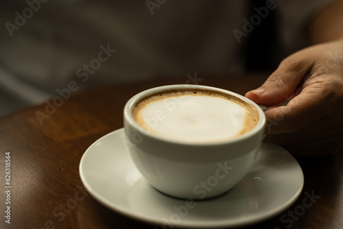 Closeup hand holding white cup of hot coffee latte on wood background in restaurant.Best of menu in the coffee shop.Soft focus.