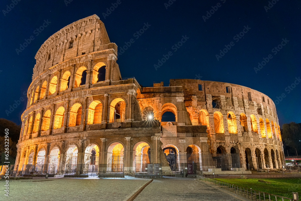 Nighttime photo of the Roman Colosseum. The structure is lit up with a warm orange glow set agains a cobalt blue sky. No people are visible.