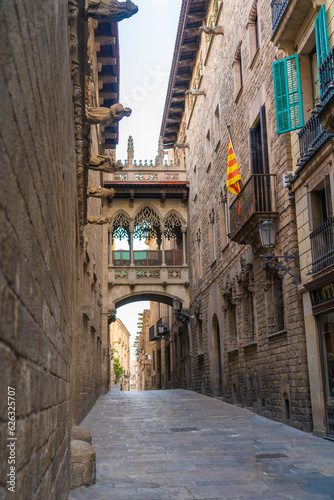 Daytime photo of the Pont del Bisbe or Bishop Street Bridge, in the Gothic Quarter of Barcelona, Spain. The Catalan flag is hanging from a balcony next to turquoise window shutters.