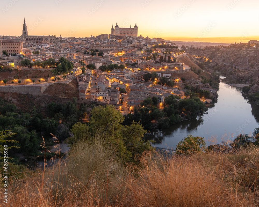 Sunrise photo of the medieval walled city of Toledo, Spain. Lights illuminate the city. Reflections of trees in the Tagus River.