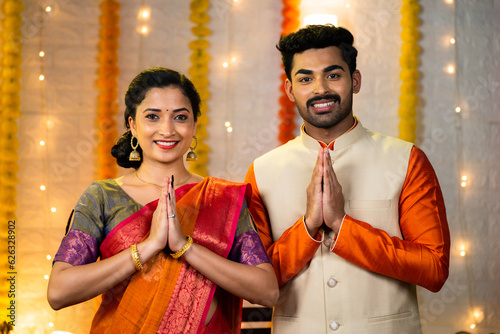 Quadro su tela Happy young indian couple in traditional ethnic clothing saying namaste or welcome gesture by looking camera during festival celebration at home - concept of greeting, wishes and togetherness
