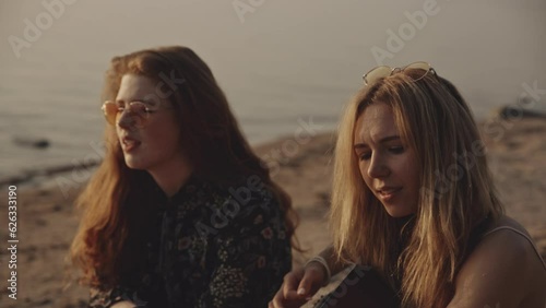 Redhead and blonde hipster girl sitting on the beach singing and playing the guitar during sunset. Vacation at sea with friends. Enjoying isolation together. Concets of friendship. travel, vacation. photo