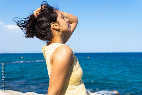 Young ethnic woman sitting on beach in daylight with eyes closed near blurred seawater