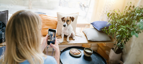 Woman taking photo of cute dog influencer. Social media content creator. Taking photo with cell phone in pet friendly cafe with wooden bench and plants. Long horizontal banner size