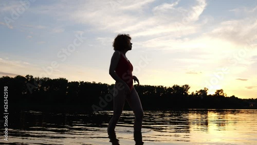 Young red-haired woman in a red swimsuit posing near the river. Slow motion motion woman swimsuit in the evening at sunset