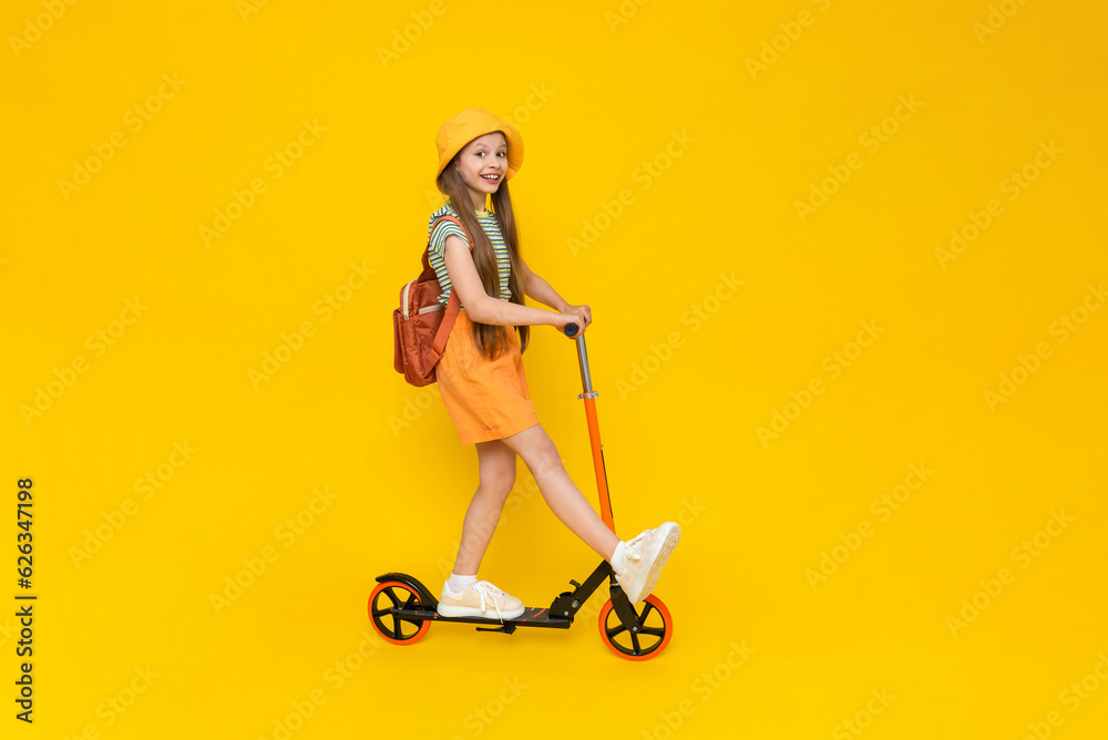 A teenage girl rides a scooter in shorts, a panama hat and with a backpack. Summer sports and active recreation for children. Yellow isolated background. A teenage girl rides a scooter in shorts, a panama hat and with a backpack. Summer sports and active recreation for children. Yellow isolated background.