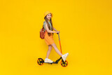 A teenage girl rides a scooter in shorts, a panama hat and with a backpack. Summer sports and active recreation for children. Yellow isolated background.