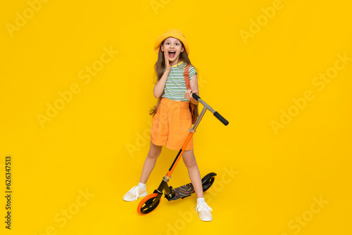 A full-length young girl in a hat and shorts is standing next to a scooter and enjoying a summer vacation. Active city recreation for children. Yellow isolated background.