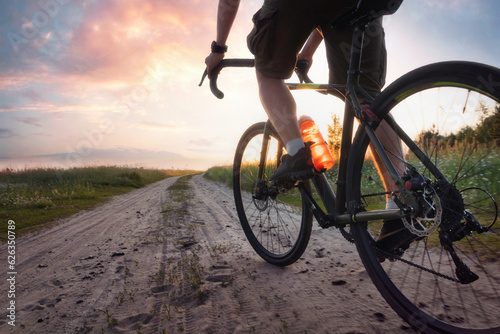 Fototapeta Naklejka Na Ścianę i Meble -  Man riding a gravel bicycle on the trail at sunset. Colorful landscape with cyclist, bike, field, green grass, dramatic sky with bright sunlight. Sport and travel. Low angle view.