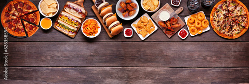 Junk food top border. Pizza, hot dogs, hamburgers, chicken wings and salty snacks. Above view over a dark wood banner background with copy space.