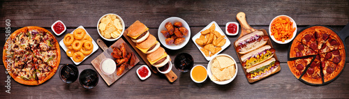 Junk food table scene. Pizza, hamburgers, chicken wings, hot dogs and salty snacks. Top down view over a dark wood banner background.