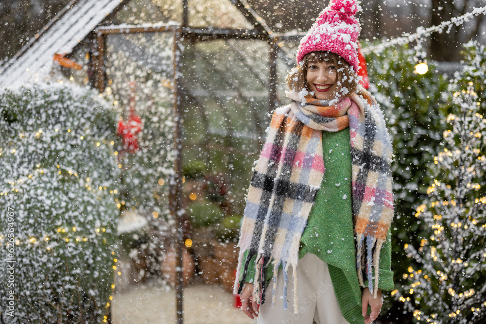 Portrait of a young woman in hat and scarf enjoying snowfall at beautifully decorated backyard. Concept of winter holidays and festive mood