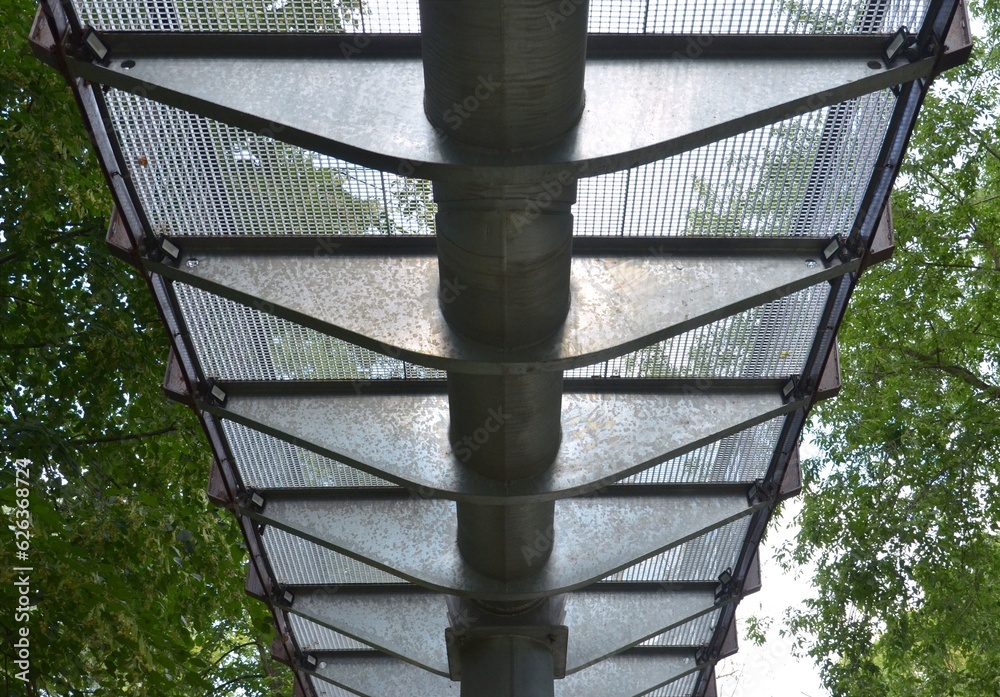 Obraz premium Metal pedestrian bridge (footbridge) in a park surrounded by green trees, close up bottom view