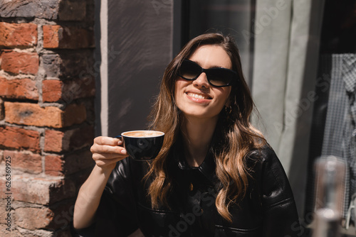 Laughing blonde girl with coffee in coffee shop terrace. Lovable lady sitting near window and holding cup of coffee and look happy. Woman drink, hold coffee mug, cappuccino foam on the lip.
