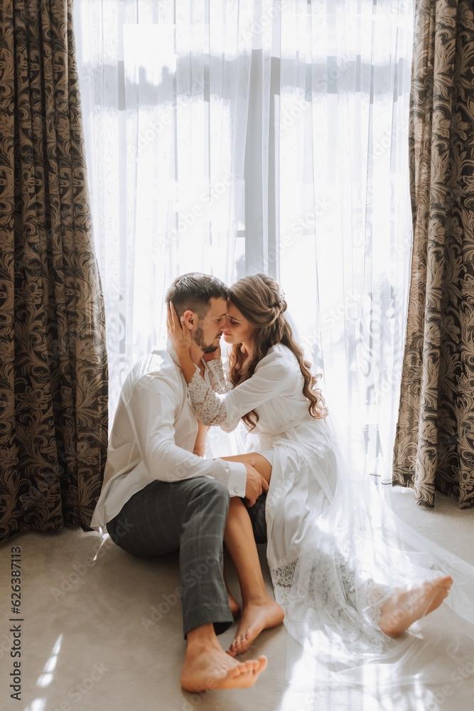 Beautiful, sexy bride in a white robe, groom in a white shirt hugs the bride against the background of the window in the hotel room. Wedding portrait of newlyweds in love. Vertical photo. Beautiful, sexy bride in a white robe, groom in a white shirt hugs the bride against the background of the window in the hotel room. Wedding portrait of newlyweds in love. Vertical photo.