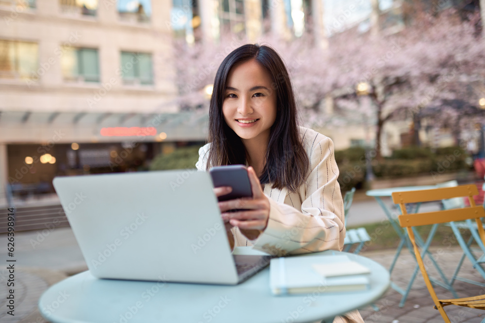 Young smiling happy Asian business woman professional or student sitting outdoors on city street at cafe table with cellphone device holding smartphone using laptop and mobile cell phone technology.