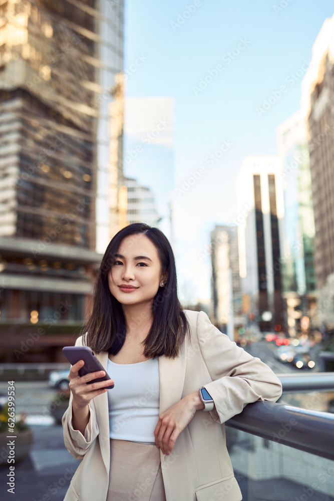 Fototapeta premium Young pretty Asian business woman office professional with cellphone in hands standing on big city urban street holding mobile cell phone, using digital apps on smartphone device, vertical.