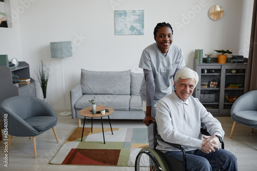 Portrait of white haired senior man in wheelchair smiling at camera indoors with young female nurse assisting, copy space