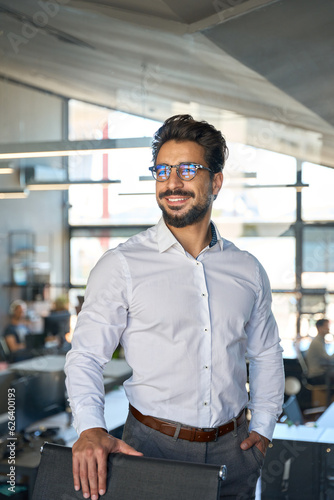 Smiling confident young Latin business man standing in office looking away. Happy Hispanic male manager entrepreneur, professional ceo executive wearing glasses thinking of financial goals, vertical.