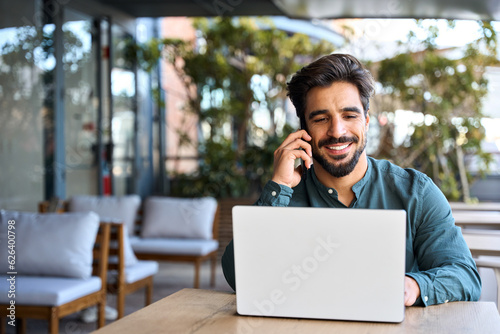 Happy young Latin business man talking on mobile phone working on laptop outdoors. Smiling professional businessman making call on cell using computer communicating with client in city cafe.