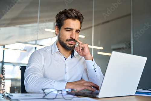 Serious busy young Latin professional business man company ceo executive manager investor looking at laptop pc computer sitting in office thinking of investment plan and financial strategy risks.