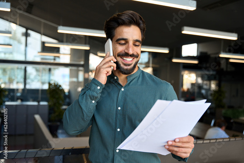 Canvas Print Smiling happy young bearded Latin professional business man executive holding documents and cell phone making mobile call at work on cellphone consulting client standing in modern office