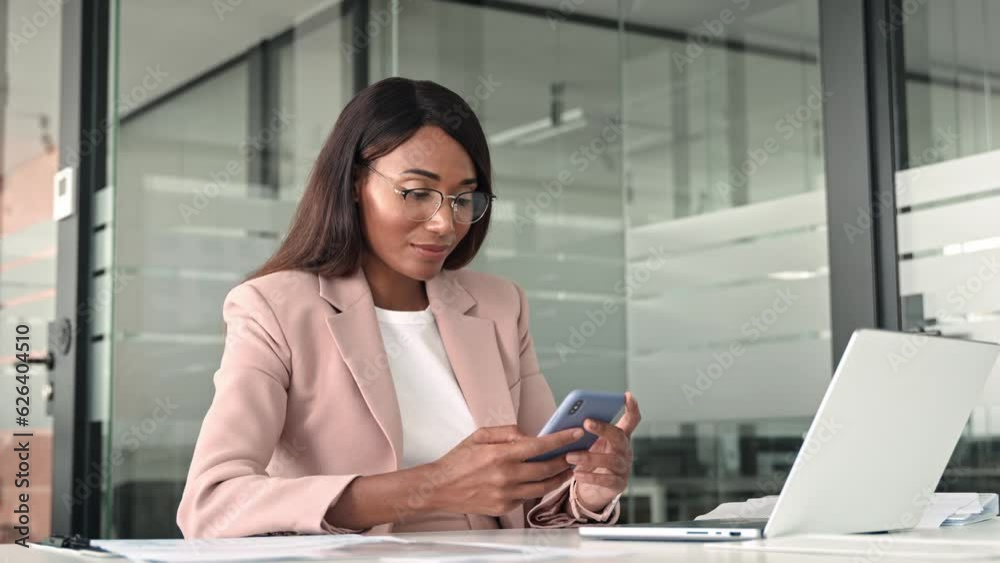 Young busy professional African American business woman executive manager holding cellphone, using mobile device looking at cell phone technology sitting at desk in office working on laptop.