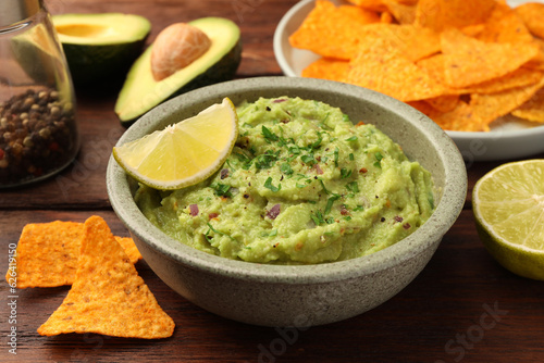 Bowl of delicious guacamole, lime and nachos chips on wooden table, closeup