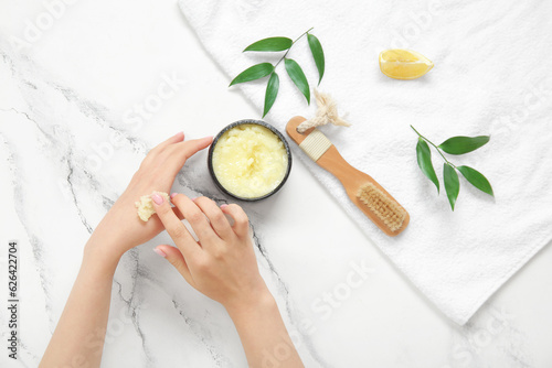 Female hands with bowl of lemon body scrub, massage brush and towel on grunge white background