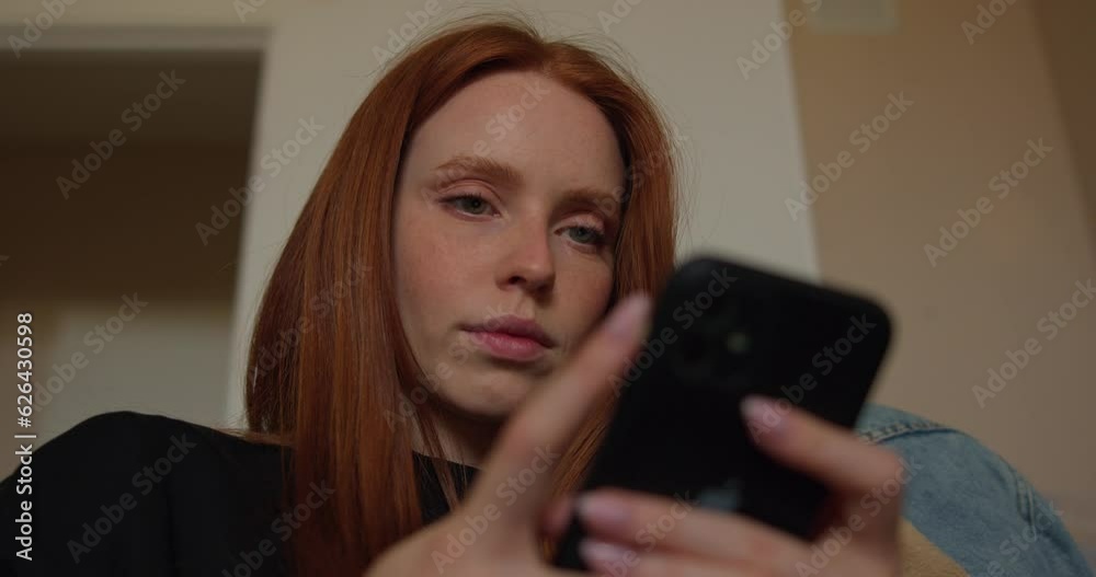 serious red-haired young woman holding smart phone looking at cellphone screen using mobile apps for shopping online playing games chatting in social media at home Technology, gadget addiction serious red-haired young woman holding smart phone looking at cellphone screen using mobile apps for shopping online playing games chatting in social media at home Technology, gadget addiction