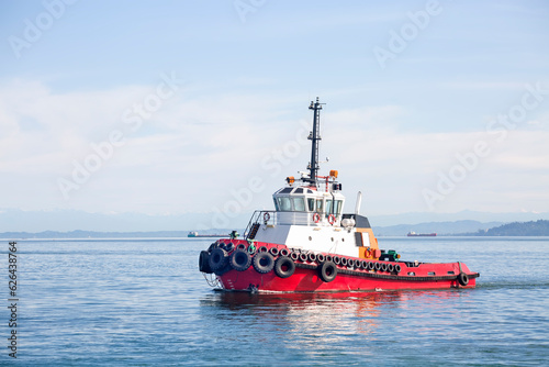 A red towboat with a white wheelhouse sailing along the hilly coast in the early morning