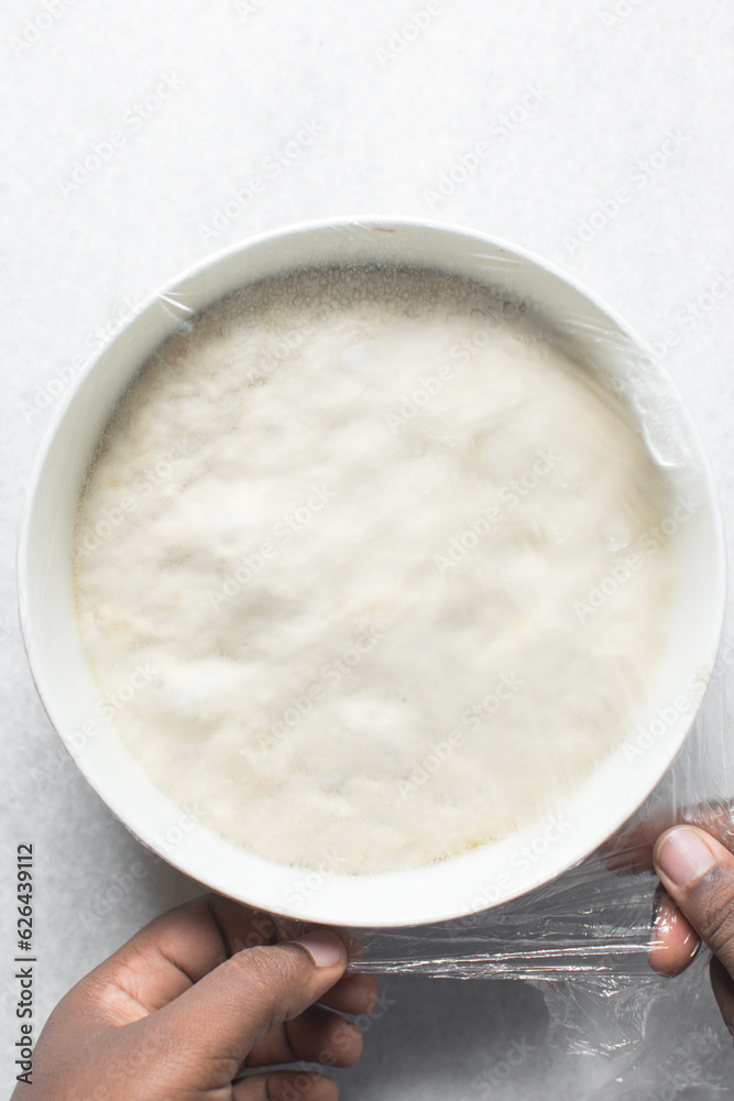 Plastic wrap covered bread dough that has doubled in size , dough that has fermented, proofed dough in a white bowl , process of making focaccia Plastic wrap covered bread dough that has doubled in size , dough that has fermented, proofed dough in a white bowl , process of making focaccia