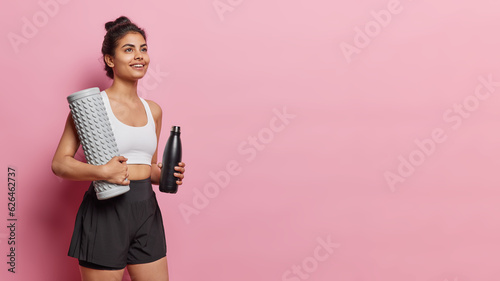 Fototapeta Naklejka Na Ścianę i Meble -  Horizontal shot of happy dreamy sporty woman has dreamy expression smiles and being deep in thoughts holds massage roller and sport bottle wears white top and black shorts isolated on pink background